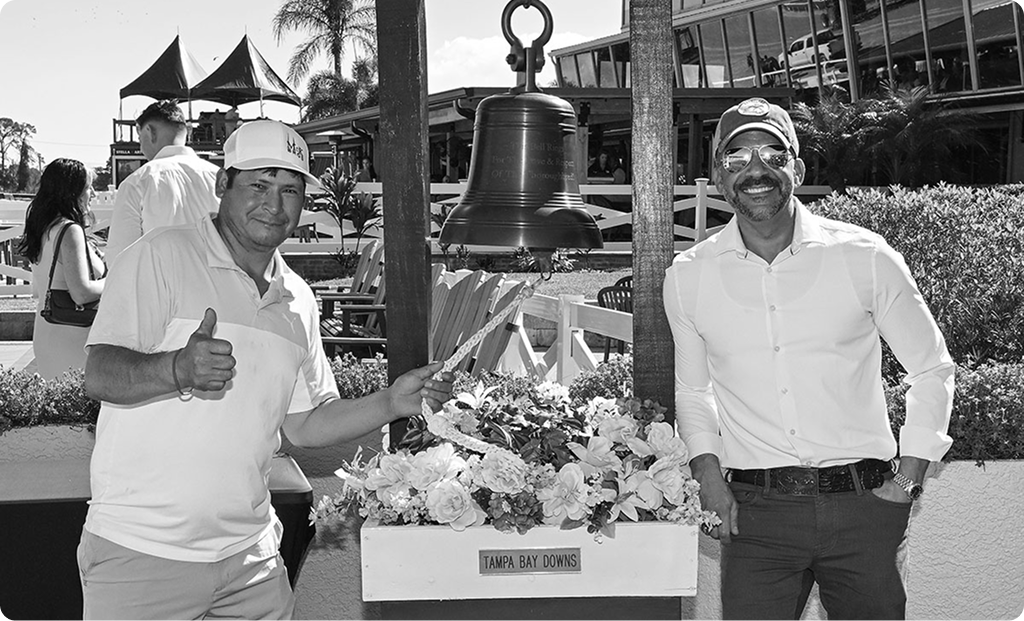 A smiling member of the Horcery team stands on the right, wearing sunglasses and a cap, next to the Tampa Bay Downs victory bell. On the left, another man gives a thumbs-up, both posing in front of a floral arrangement and event backdrop.