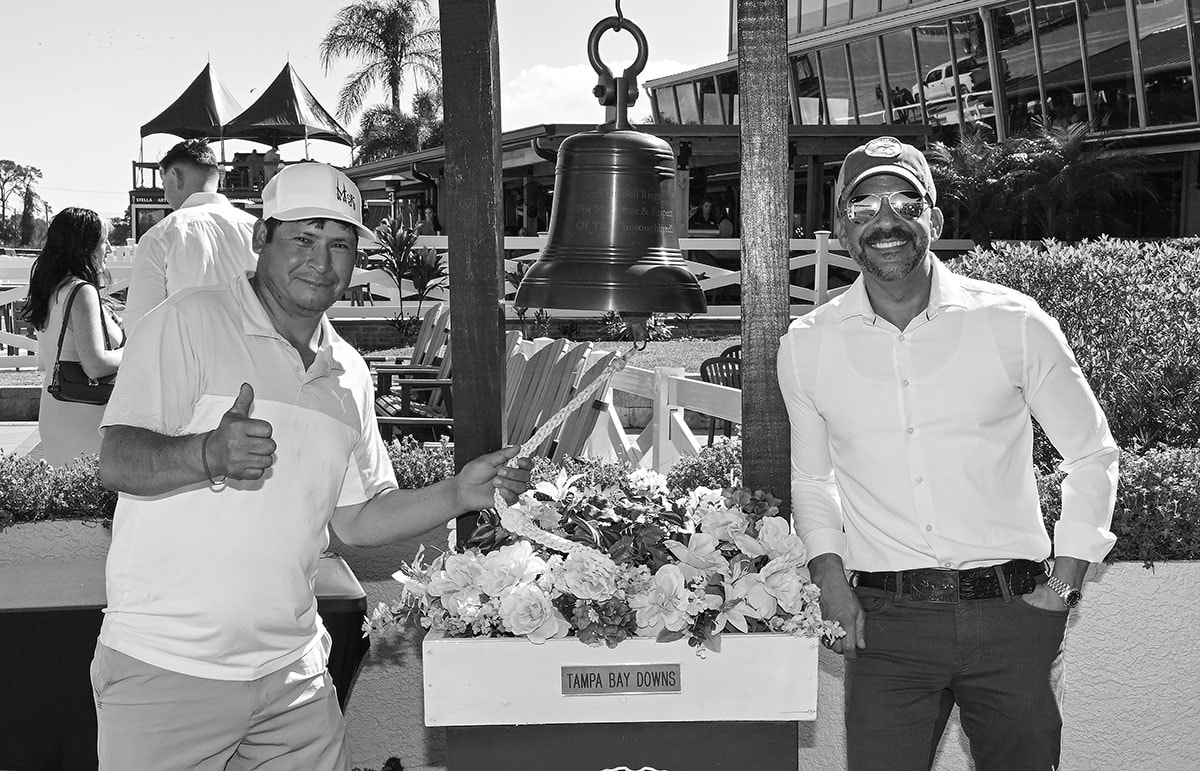 A smiling member of the Horcery team stands on the right, wearing sunglasses and a cap, next to the Tampa Bay Downs victory bell. On the left, another man gives a thumbs-up, both posing in front of a floral arrangement and event backdrop.