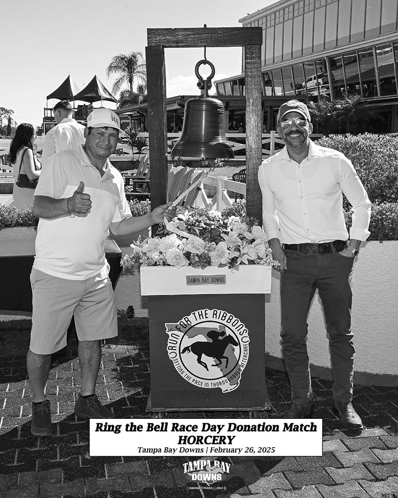 A smiling member of the Horcery team stands on the right, wearing sunglasses and a cap, next to the Tampa Bay Downs victory bell. On the left, another man gives a thumbs-up, both posing in front of a floral arrangement and event backdrop.