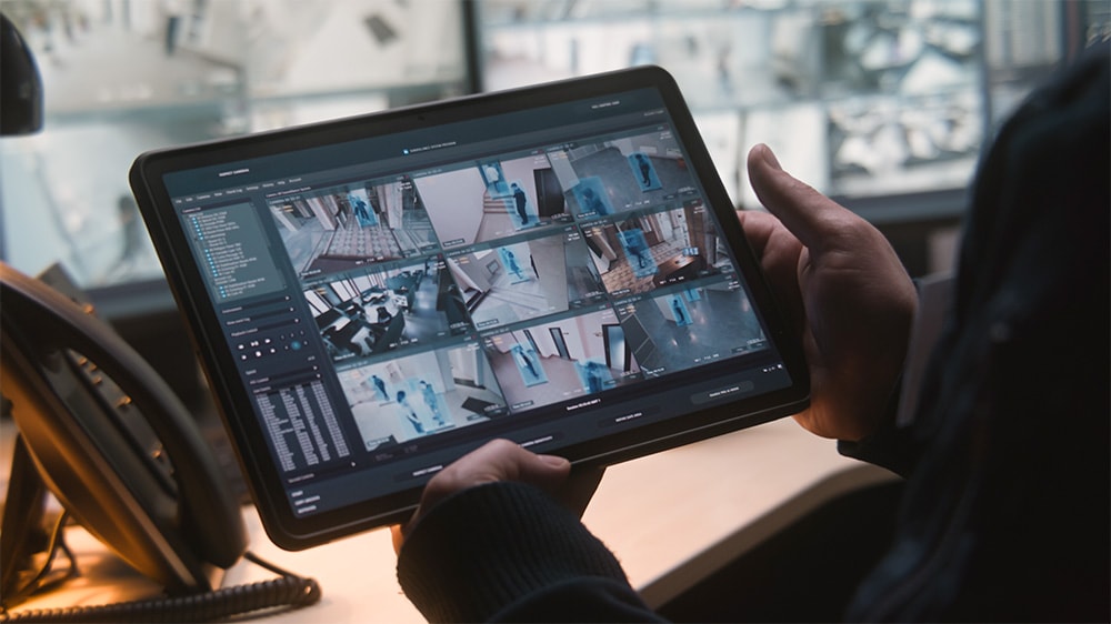 A person holds a tablet displaying a multi-camera surveillance interface with nine video feeds showing indoor activity. Each feed includes motion detection highlights in blue. The setting appears to be a modern security or monitoring control room with blurred screens in the background.
